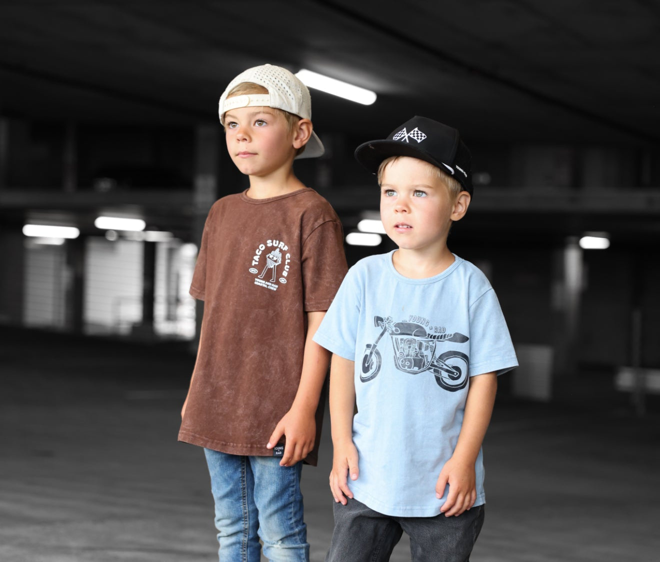 Two children standing in an indoor setting wearing cool graphic t-shirts and caps with motorcycles and surf style.